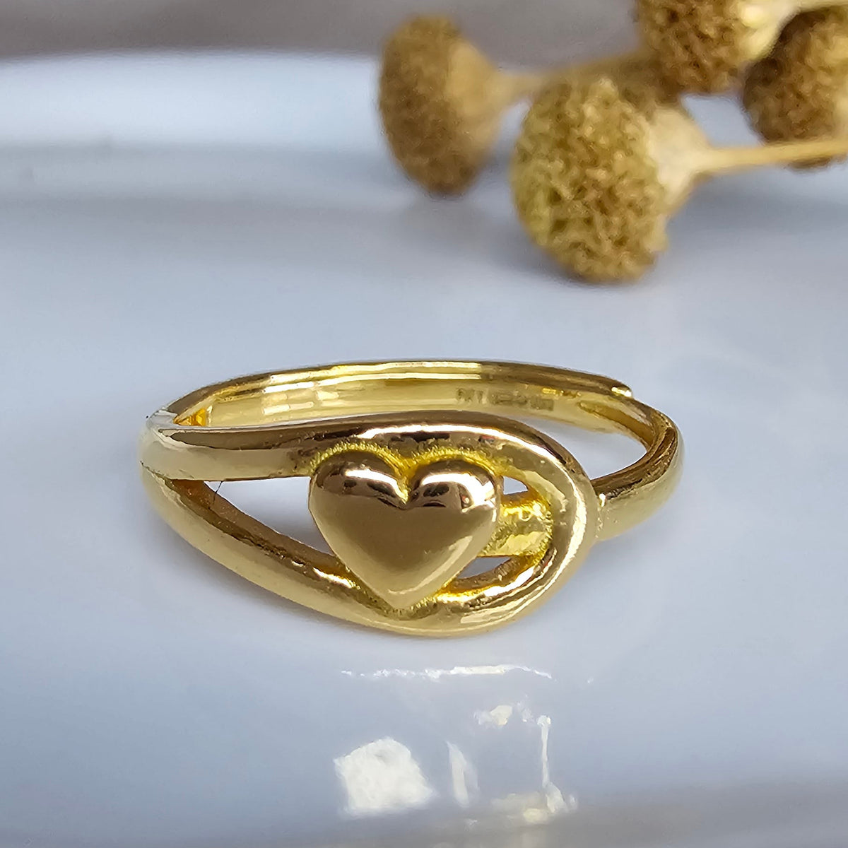 A solid gold ring with a heart-shaped center and a curved wrap-around band, displayed on a glossy white surface with dried flowers in the background.