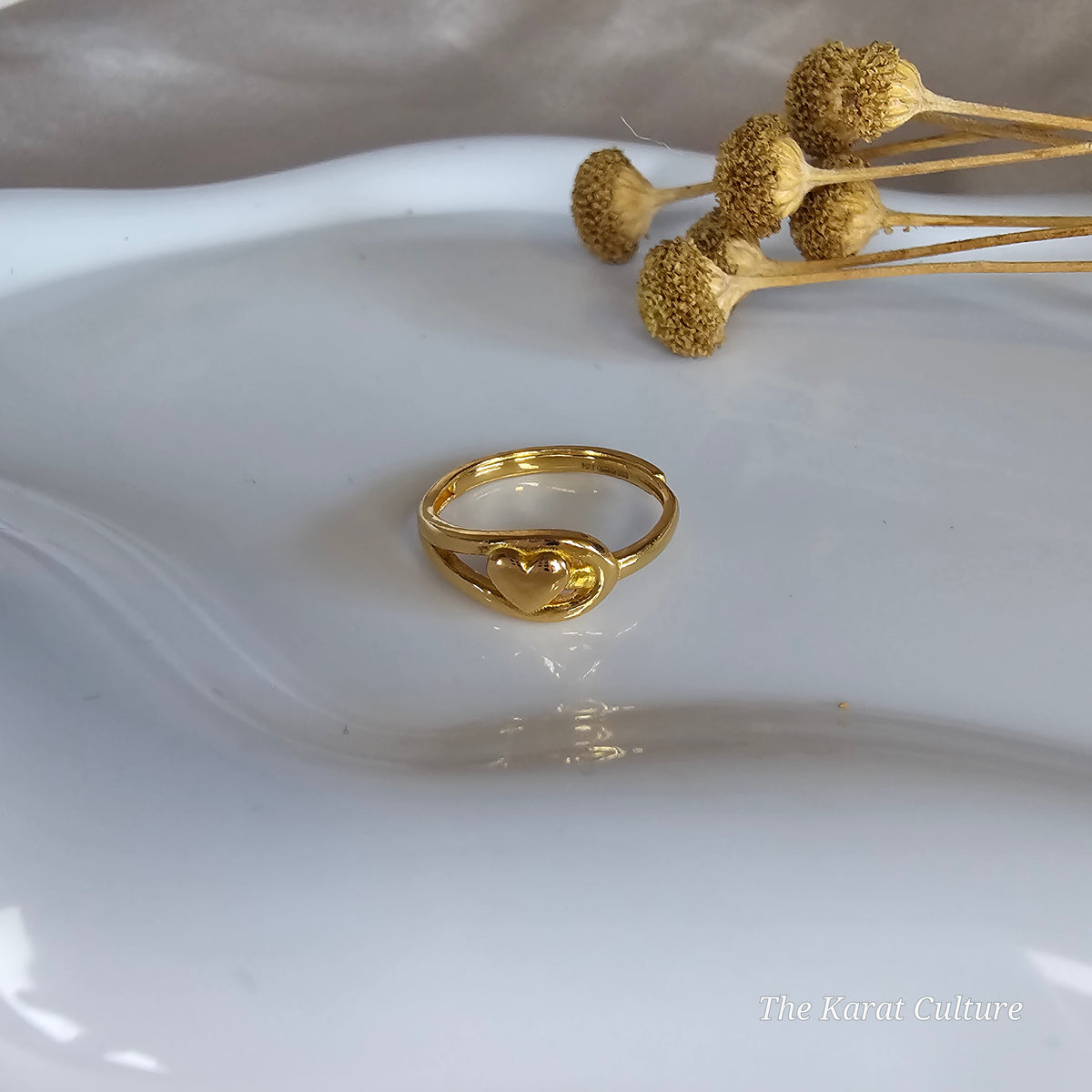 A gold ring with a heart-shaped knot design, displayed on a white ceramic dish with dried flower stems in the background.