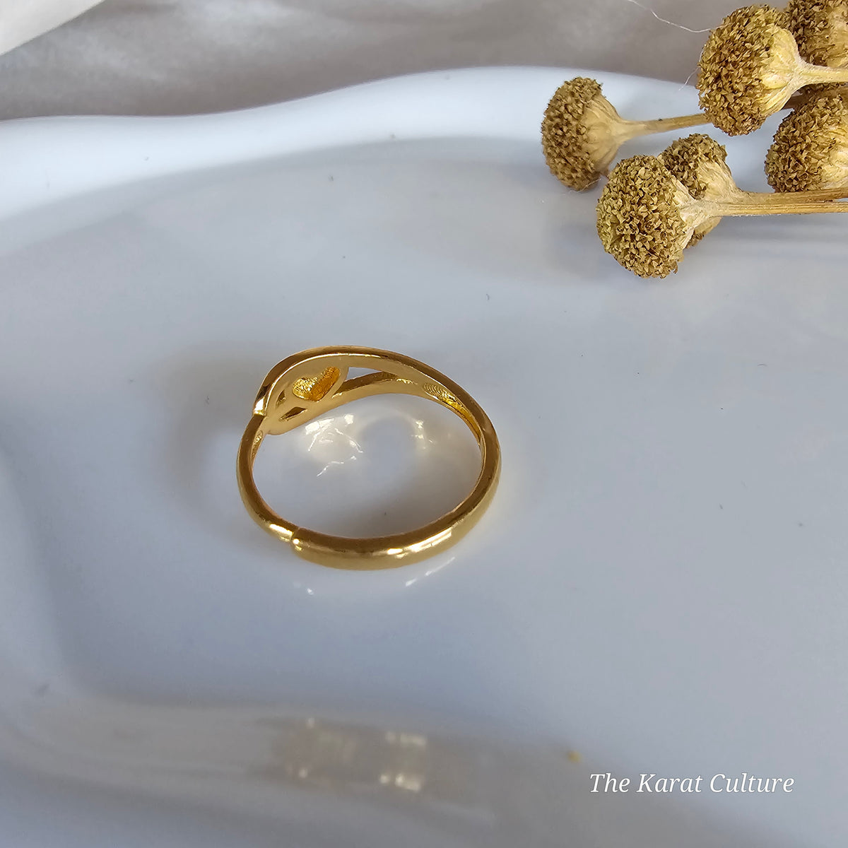 A solid gold  ring with a polished finish, displayed on a white ceramic dish with dried flower stems in the background.